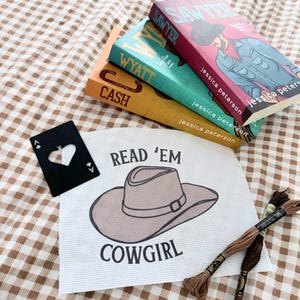 May include: Three books titled "Sawyer", "Wyatt", and "Cash" with author Jessica Peterson, stacked on a brown and white checkered surface. A black ace of hearts playing card and a white fabric with a cowboy hat graphic and the text "Read 'Em Cowgirl" are also present.