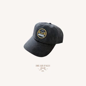 May include: A dark gray baseball cap with a curved brim. The cap features a circular patch with the text "RENEGADES" and a graphic of a bridge and football. The hat is made of a soft material.
