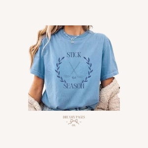 Op de afbeelding: Lichtblauw T-shirt met de tekst "STICK SEASON" in een cirkelvormig ontwerp met gekruiste hockeysticks en een puck, omringd door een lauwerkrans. Het T-shirt heeft korte mouwen en is casual.