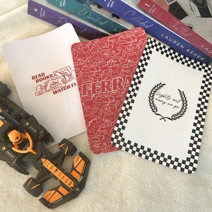 May include: An assortment of items related to Formula 1 racing. Includes books from the "Dirty Air Series", a toy race car, and several cards with racing-themed designs. One card reads "Read Books & Watch F1". Another card has the word "FERRARI".