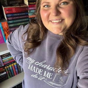 May include: A lilac-colored sweatshirt with the text "my characters MADE ME do it" in white script. The design includes a laptop graphic with a person sitting inside. Bookshelves filled with books are in the background.