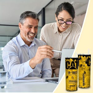 Op de afbeelding: Twee cilindrische bekers met het CAT-logo en de tekst "1R-1807 Engine Oil Filter" in zwart en geel. De bekers hebben een verweerd, industrieel ontwerp. Twee mensen lachen en kijken naar een laptop.