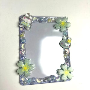 May include: A rectangular, clear frame with a decorative border. The border is embellished with small, iridescent beads, crescent moons, and cartoon bunny and flower accents. The frame has a reflective surface.