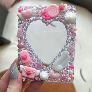 May include: A clear, heart-shaped frame decorated with pink and white embellishments. The frame is adorned with candy-themed charms, including a lollipop, ice cream, and hearts. The frame is surrounded by iridescent beads.