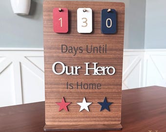 Puede incluir: Calendario de cuenta regresiva de madera con el texto "Days Until Our Hero Is Home". Cuenta con detalles en rojo, blanco y azul, incluyendo los números "130" y tres estrellas. El diseño es patriótico y decorativo.