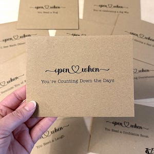 May include: A collection of kraft paper cards with the phrase "open when" and various prompts printed in black script. One card reads "You're Counting Down the Days." Other cards include phrases like "You Need a Hug" and "You Need a Confidence Boost."