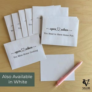 May include: White paper envelopes with the text "open when" and various phrases, including "You Miss Home Cooking" and "You Need to Have Some Fun." A pink pen and a blank white card are also present. The phrase "Also Available in White" is displayed.