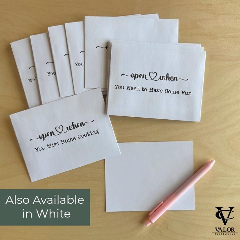May include: White paper envelopes with the text "open when" and various phrases, including "You Miss Home Cooking" and "You Need to Have Some Fun." A pink pen and a blank white card are also present. The phrase "Also Available in White" is displayed.