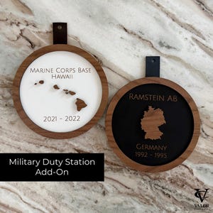 May include: Two wooden military duty station add-ons. One features the Hawaii islands and text "MARINE CORPS BASE HAWAII 2021-2022". The other shows Germany with "RAMSTEIN AB GERMANY 1992-1995". Both have a dark brown leather strap.