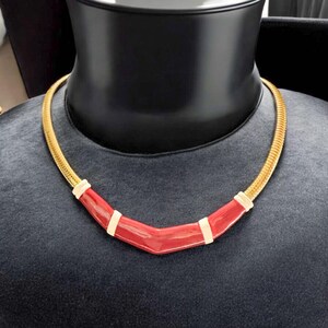 May include: A gold chain necklace with a red and white enamel pendant. The pendant is shaped like a curved line.