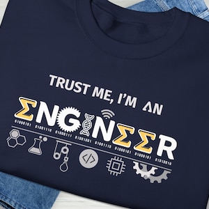 Könnte beinhalten: Marineblaues T-Shirt mit dem weißen Text "TRUST ME, I'M AN ENGINEER". Das Wort "ENGINEER" ist stilisiert mit gelben und weißen Akzenten und Binärcode. Enthält technische Symbole.