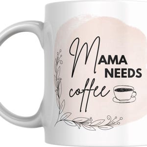 May include: White ceramic coffee mug with a pink watercolor background and black text that reads "Mama Needs Coffee". The text is surrounded by black floral illustrations.