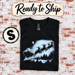 Op de afbeelding: Een zwart t-shirt met een witte afbeelding van de Kerstman in een arrenslee getrokken door rendieren die over een bos vliegen. De tekst Ready to Ship staat bovenaan en een ronde badge met de letter S staat links op het shirt.