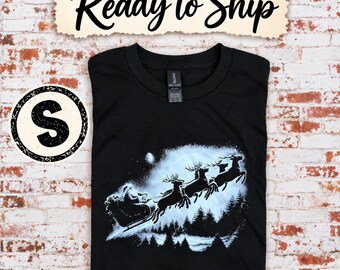 Puede incluir: Una camiseta negra con un gráfico blanco de Santa Claus en un trineo tirado por renos volando sobre un bosque. El texto Ready to Ship está en la parte superior y una insignia circular con la letra S está en el lado izquierdo.
