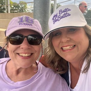 May include: A light purple baseball cap with the number 18 and the word mama embroidered in purple, and a white baseball cap with the text Hey Batta Batta and a small baseball graphic embroidered in purple on the front panel.