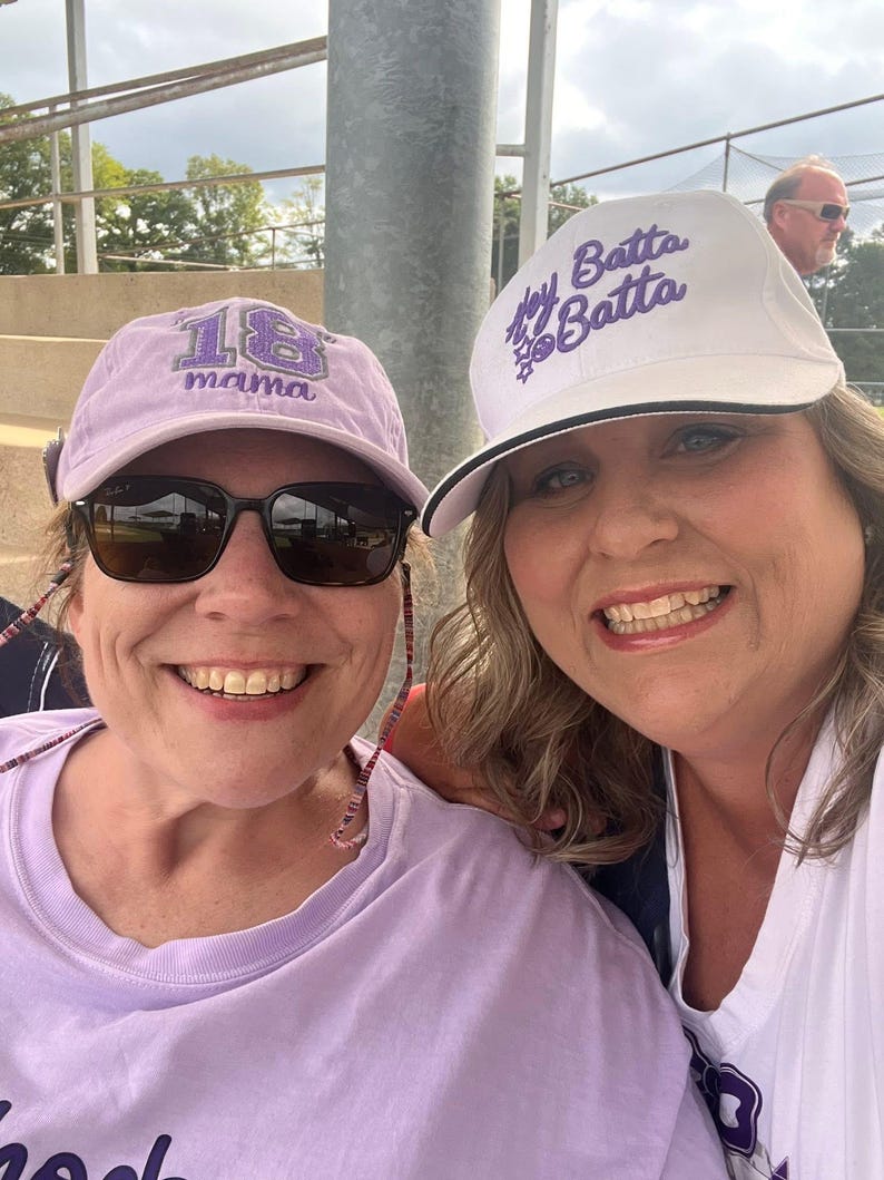 May include: A light purple baseball cap with the number 18 and the word mama embroidered in purple, and a white baseball cap with the text Hey Batta Batta and a small baseball graphic embroidered in purple on the front panel.