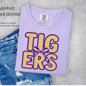 May include: A light purple short-sleeve t-shirt with a crew neckline and a Comfort Colors brand label. The shirt features the text TIGERS in yellow vertical stripes with a purple scalloped outline and a purple bow graphic centered on the word.
