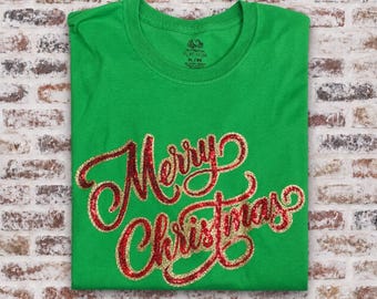Puede incluir: Una camiseta verde brillante de manga corta con el texto Merry Christmas en letras cursivas de purpurina roja y dorada en el pecho. La prenda tiene cuello redondo y una etiqueta Fruit of the Loom Platinum impresa en el cuello interior.
