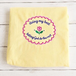 May include: A pale yellow t-shirt featuring a central embroidered design. The embroidery shows a pink tulip with green leaves inside a scalloped pink border, with the text "doing my best" above and "letting God do the rest" below in blue.