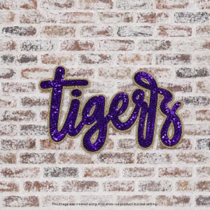 May include: A purple sequin patch in a cursive script font that spells out the word tigers. The letters are outlined in a gold metallic thread, creating a bold and reflective appearance against the textured brick background.