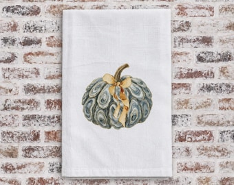 May include: A white rectangular kitchen towel featuring a central illustration of a pumpkin constructed from numerous grey and blue oyster shells. A light tan ribbon is tied in a bow around the stem of the shell pumpkin design.