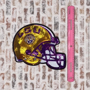 May include: A sequined football helmet patch in gold and purple featuring a tiger logo and the text LSU. The patch is positioned next to a pink ruler showing a height of approximately 4 inches for the helmet design.