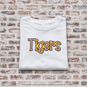 May include: A white crew neck t-shirt featuring the word Tigers embroidered in yellow thread with a dark purple outline across the center of the chest. The garment has short sleeves and a standard fit, presented in a clean, folded arrangement.