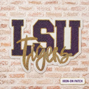 May include: An iron-on patch featuring the letters LSU in purple rhinestones with a white border, layered with the word Tigers in gold rhinestones in a cursive script font. The patch has a white backing and a rectangular label reading IRON-ON PATCH.