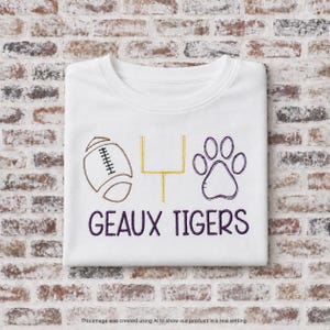 Op de afbeelding: Een wit t-shirt met een geborduurd ontwerp van een bruine football, een gouden doelpaal en een paarse pootafdruk. Onder de iconen staat de tekst GEAUX TIGERS geborduurd in paarse letters op de voorzijde van het t-shirt.