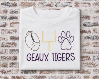 May include: A white t-shirt featuring an embroidered design with a brown football, a gold goalpost, and a purple paw print. Below the icons, the text GEAUX TIGERS is embroidered in purple lettering. The design is centered on the front of the shirt.