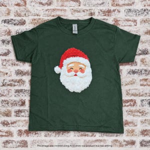 Op de afbeelding: Een donkergroen T-shirt met korte mouwen en een centrale afbeelding van een kerstmangezicht. De afbeelding toont een rode muts met een witte pompon, een witte baard, een witte snor en een perzikkleurig gezicht met gesloten ogen.