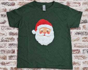 Puede incluir: Una camiseta de manga corta de color verde oscuro con un gráfico central de la cara de Santa Claus. El diseño muestra un gorro rojo con un pompón blanco, una barba blanca, un bigote blanco y una cara color melocotón con ojos cerrados.