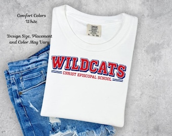 May include: A white crew neck t-shirt featuring the red and blue text WILDCATS and CHRIST EPISCOPAL SCHOOL with blue horizontal lines. The shirt has a Comfort Colors brand label at the neckline and is paired with blue denim shorts.