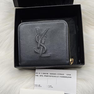 Peut inclure: Un portefeuille en cuir gris foncé avec une fermeture éclair dorée et le logo YSL en relief sur le devant, présenté dans une boîte noire avec le logo Yves Saint Laurent rive gauche. Inclut une carte d'information produit et une carte blanche.