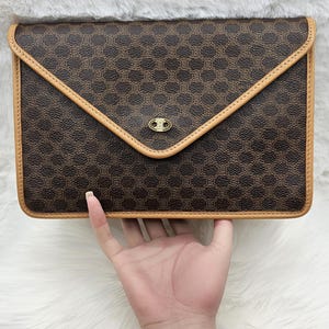 May include: A brown and black envelope-style clutch bag featuring a repeating circular monogram pattern. The bag is trimmed with light tan leather piping along the edges and includes a gold-toned metal emblem centred on the triangular flap closure.