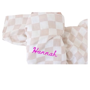 Puede incluir: Una manta de bebé a cuadros beige y blanco con el nombre "Hannah" bordado en rosa.