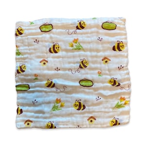 Könnte beinhalten: Ein weißer Musselin-Spucktuch für Babys mit einem sich wiederholenden Muster aus Bienen, Blumen und Häusern. Die Bienen sind braun und gelb mit schwarzen Streifen. Die Blumen sind gelb und orange. Die Häuser sind braun.