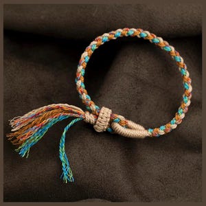 Puede incluir: Un brazalete trenzado con un borla hecha de hilos marrones, naranjas, azules y verdes. El brazalete está atado con un nudo y tiene una borla marrón y blanca.