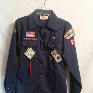 May include: A navy blue Boy Scouts of America uniform shirt with long sleeves and a button-down collar. The shirt has several patches, including a US flag patch, a "Progress Toward Ranks" patch, a "Bear" patch, and a "Bobcat" patch. The shirt also has a red bead necklace.
