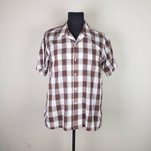 May include: A short-sleeved, button-down shirt in a brown and white checkered pattern. The shirt has a classic collar and two chest pockets. It is displayed on a black mannequin against a plain background. The shirt is a vintage style.