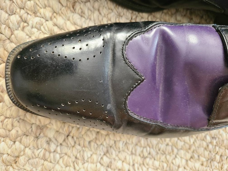 May include: Black leather oxford shoes with a purple leather panel on the toe. The shoes have a brogue design with small holes.