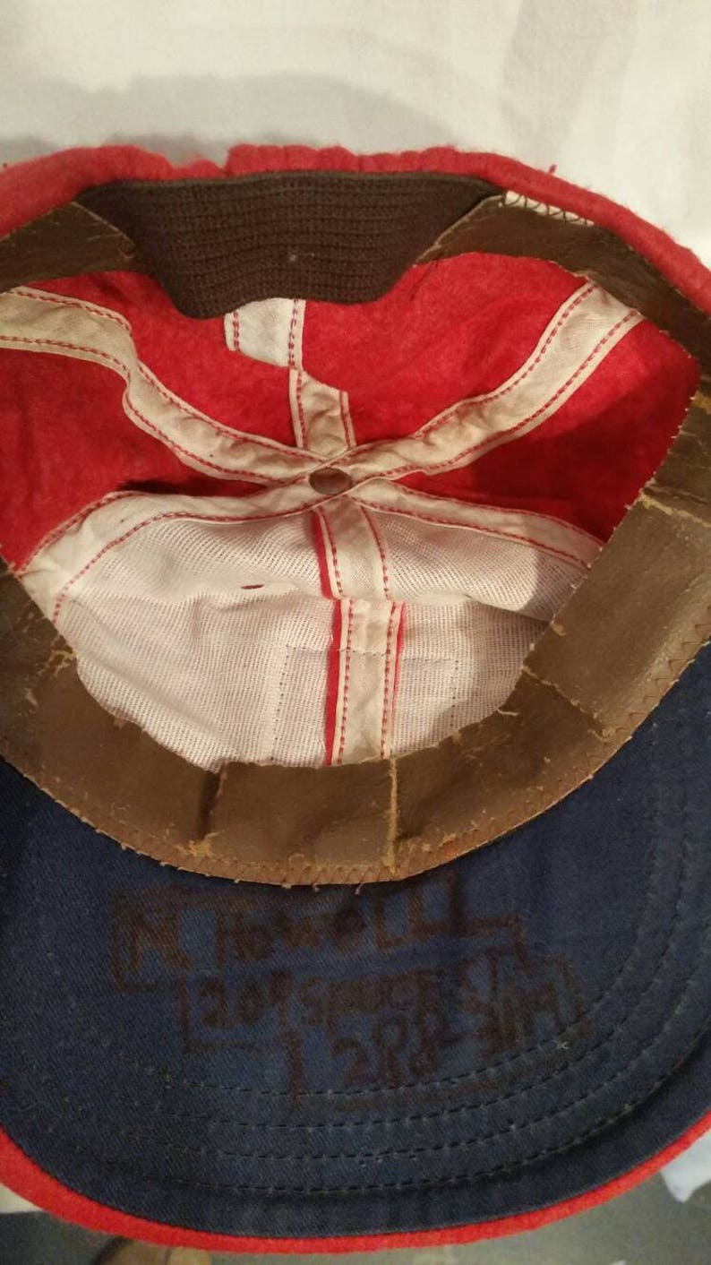 May include: Red and blue baseball cap with a brown leather band. The inside of the cap is white with red stitching. The cap has the text "FT. WORTH 1947 FIRE DEPT." written in black ink on the inside.