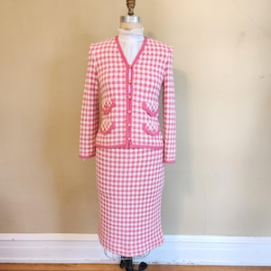 May include: A pink and white gingham patterned skirt suit with a white collared shirt underneath. The jacket has gold buttons and a scalloped trim.