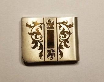 May include: A silver money clip with a gold-colored design of a stylized floral pattern.