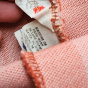 May include: Close-up of a peach-colored garment with a white tag that reads "MACHINE WASH COLD", "HAND WASH", "LIKE COLORS", "TUMBLE DRY LOW", "DO NOT BLEACH", "IRON LOW", "MADE IN ITALY" and another white tag that reads "FRONT".
