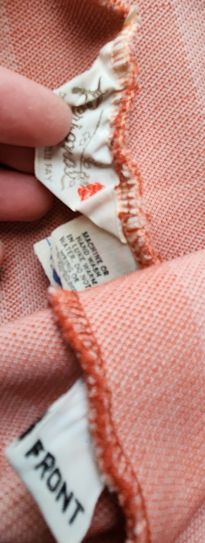 May include: Close-up of a peach-colored garment with a white tag that reads "MACHINE WASH COLD", "HAND WASH", "LIKE COLORS", "TUMBLE DRY LOW", "DO NOT BLEACH", "IRON LOW", "MADE IN ITALY" and another white tag that reads "FRONT".