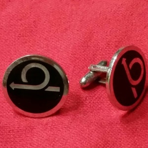 May include: A pair of silver cufflinks with black enamel. One cufflink features a white stylized letter 'P' and the other cufflink features a white stylized curved arrow.
