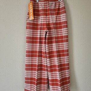 May include: Red and white plaid pants hanging on a hanger. The pants have a wide leg and are made of a woven fabric. The tag on the pants reads "PERSONAL".