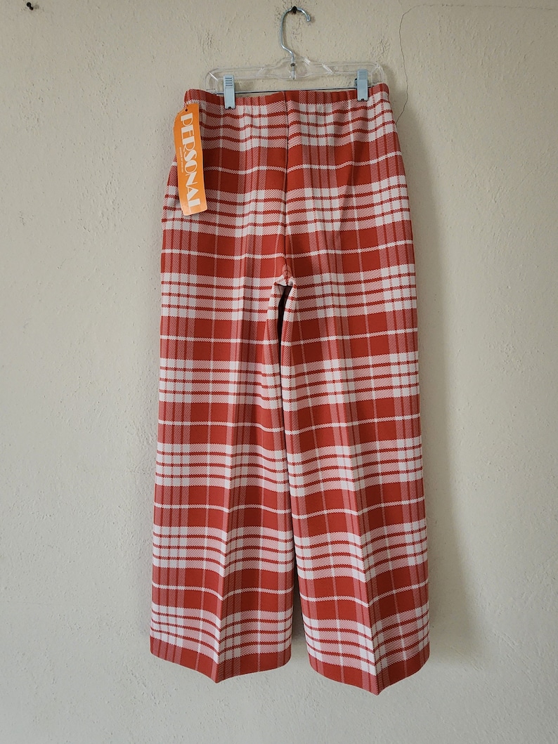 May include: Red and white plaid pants hanging on a hanger. The pants have a wide leg and are made of a woven fabric. The tag on the pants reads "PERSONAL".