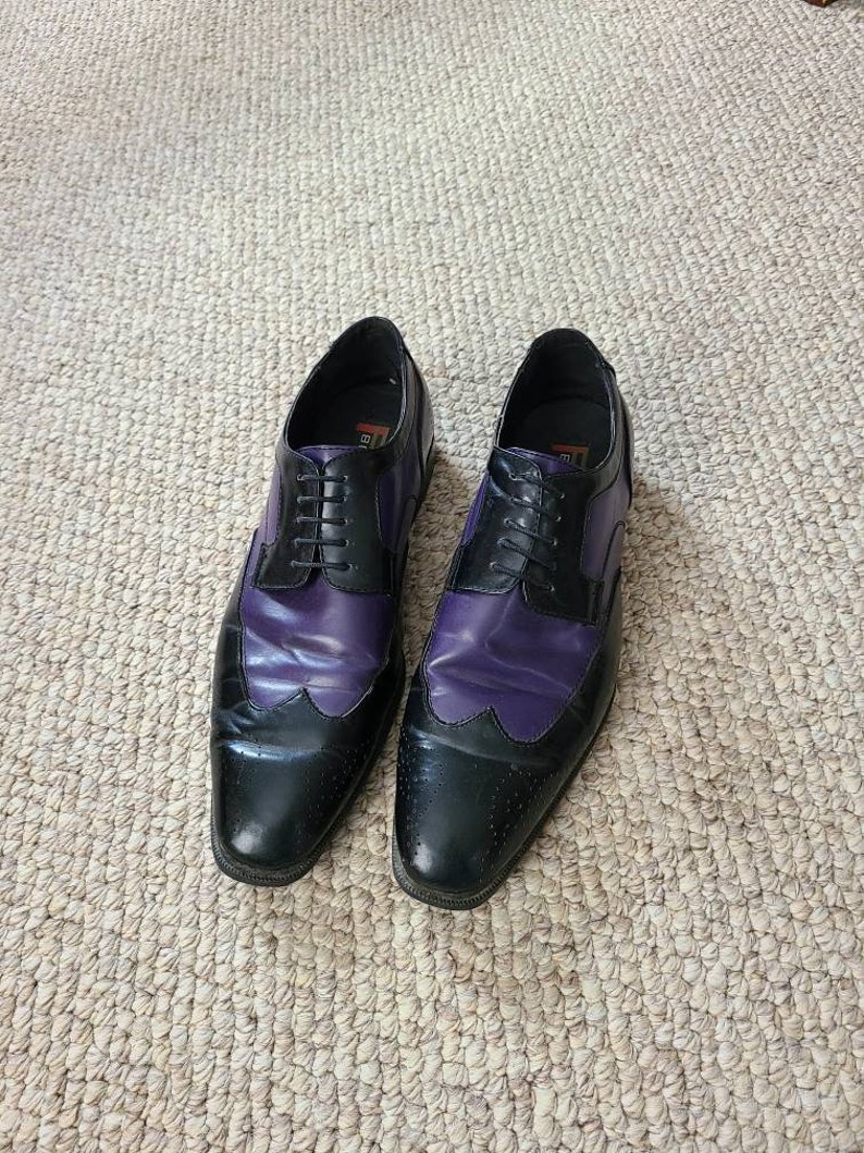 May include: A pair of black and purple leather dress shoes with lace-up closures. The shoes have a wingtip design and a pointed toe.